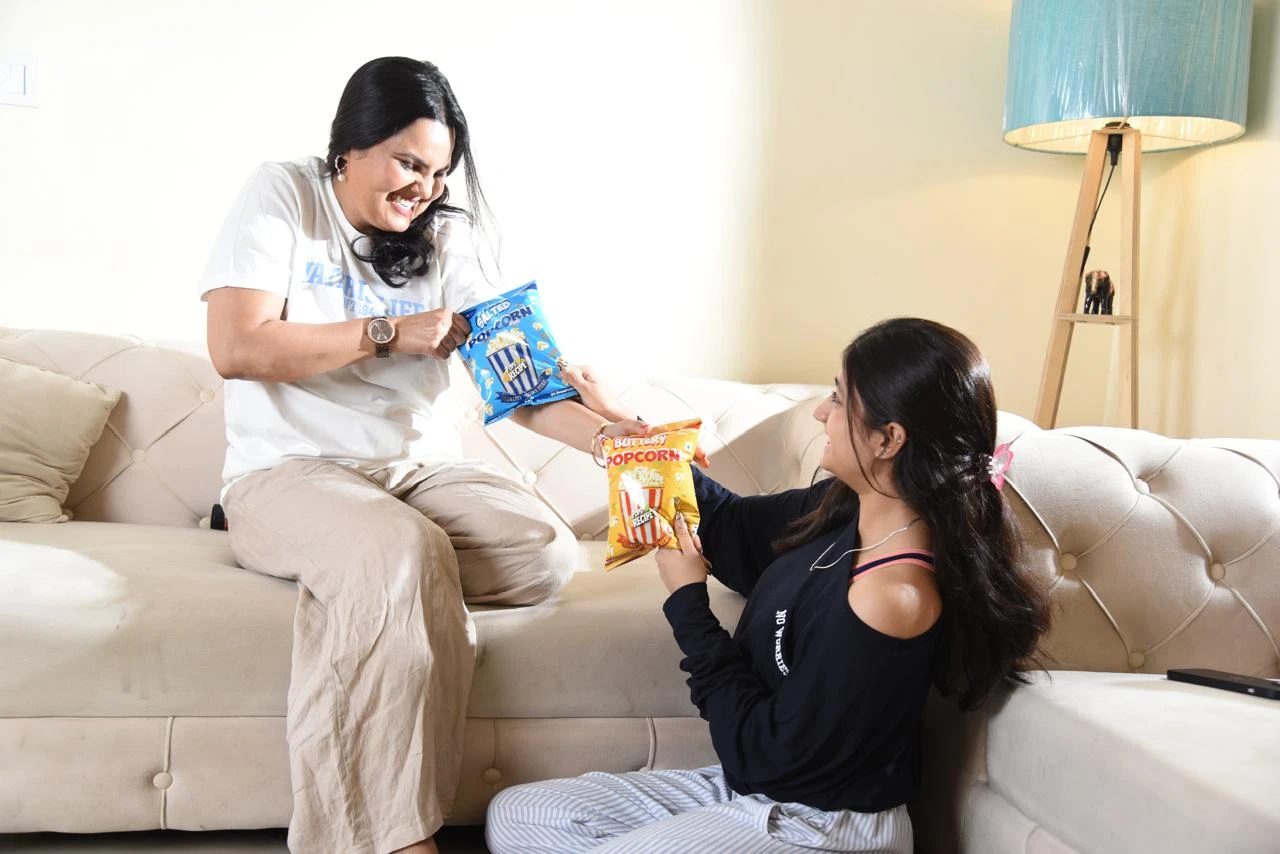 Two women laughing on a sofa as they share Pops Recipe Salted and Buttery popcorn packs